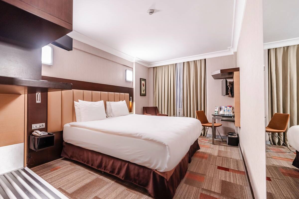 A hotel room with a large bed, a desk, a chair, a phone, and a window with curtains.