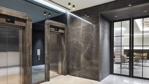 Un ascenseur moderne doté d’un écran numérique, de murs en marbre et d’une porte en verre donnant sur un espace de restauration avec des tables et des chaises.