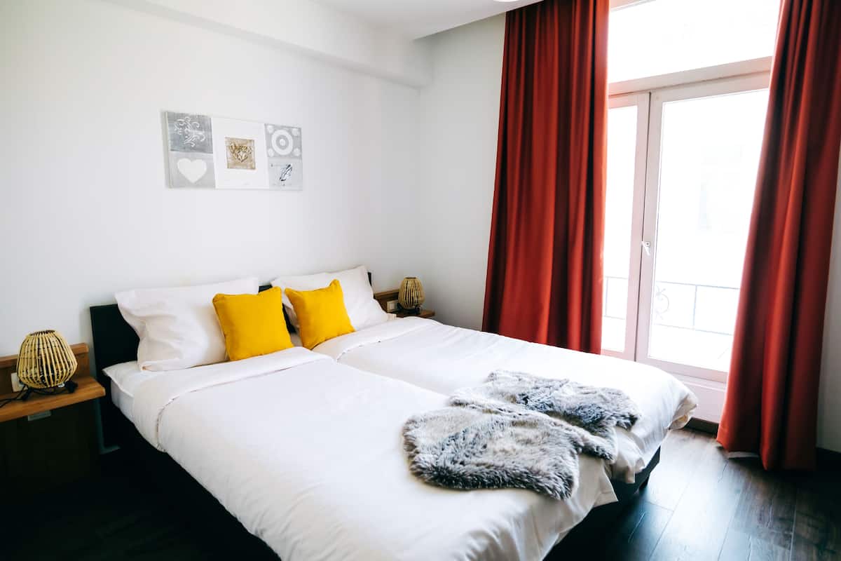 A double bed with white bedding and two pillows, one yellow and one white, in a room with a large window and red curtains.