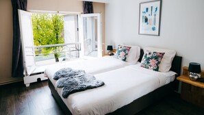 A bedroom with a large bed, a window with curtains, a nightstand, and a framed picture on the wall. - Hotel Den Grooten Wolsack (Mechelen)