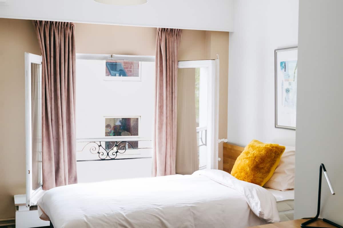 A modern bedroom with a large bed, a wooden desk, a desk lamp, and a view of the outdoors through a window with curtains.