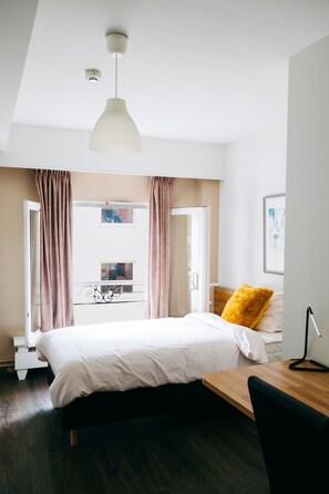 A modern bedroom with a large bed, a wooden desk, a desk lamp, and a view of the outdoors through a window with curtains. - Hotel Den Grooten Wolsack (Mechelen)