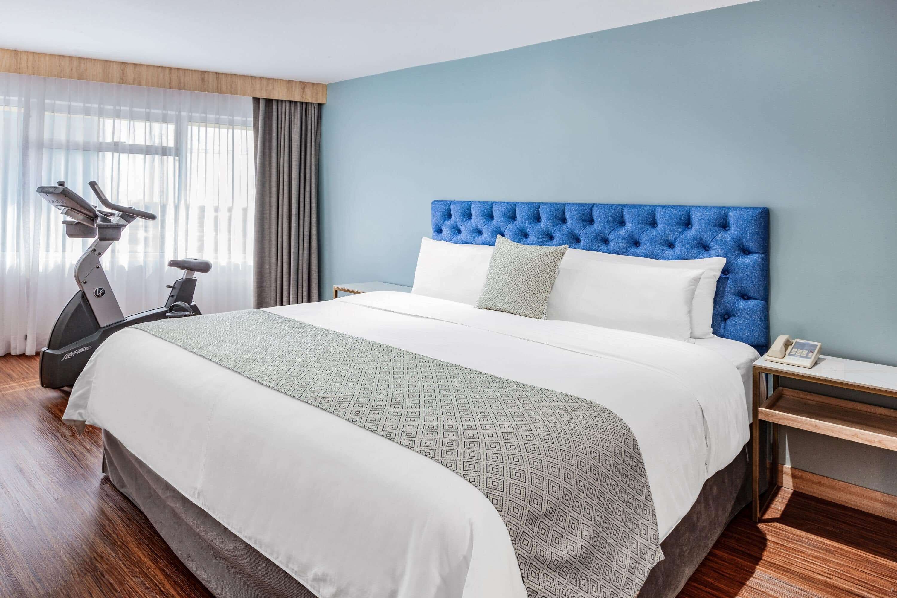 Quarto de hotel com uma cama grande, cabeceira estofada, mesa de cabeceira com telefone e uma bicicleta ergométrica.