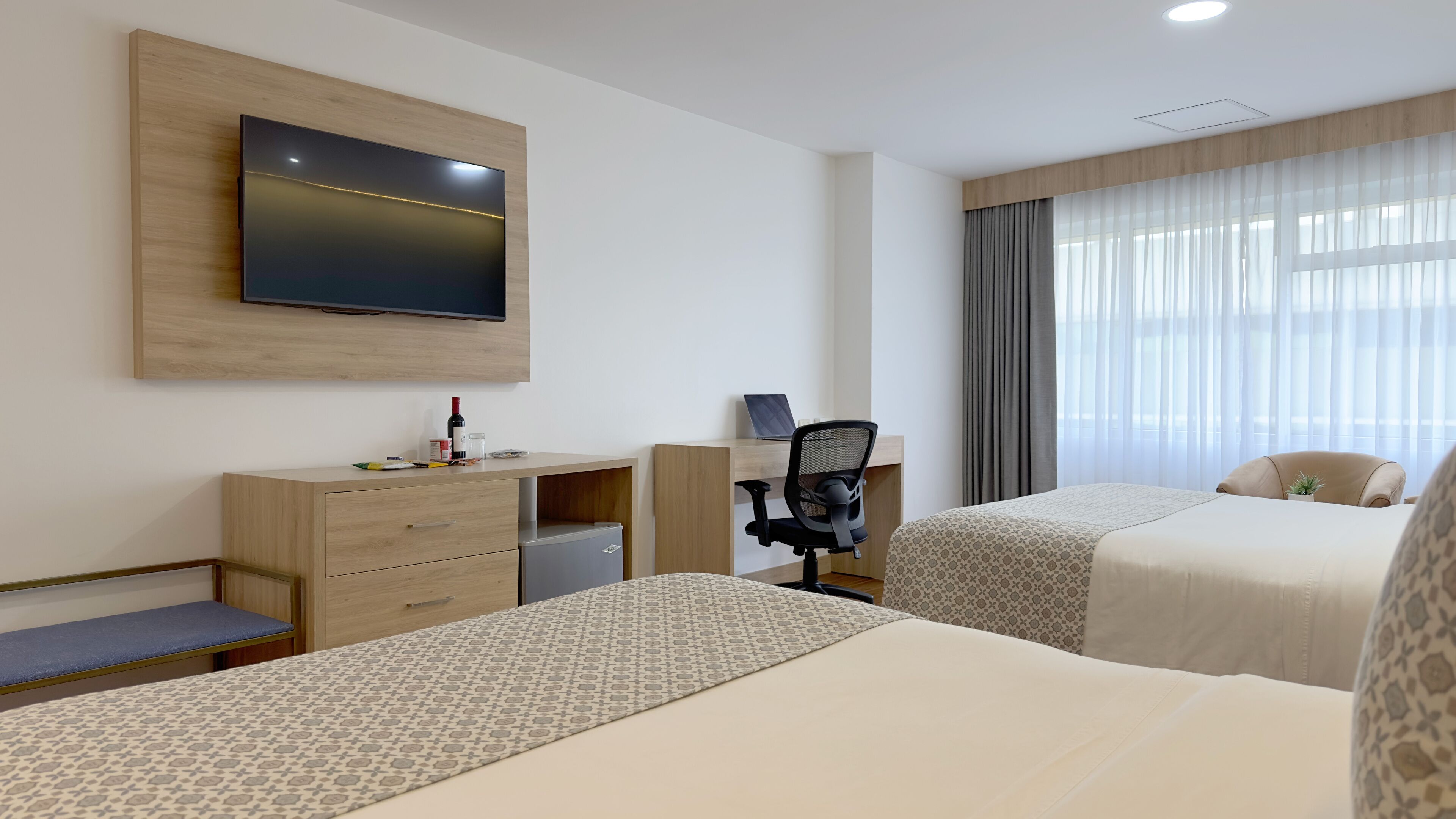 A hotel room with two beds, a desk with a chair, a television mounted on the wall, and a window with curtains.