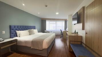 A hotel room with a large bed, a desk, a chair, a TV mounted on the wall, and a window with curtains.