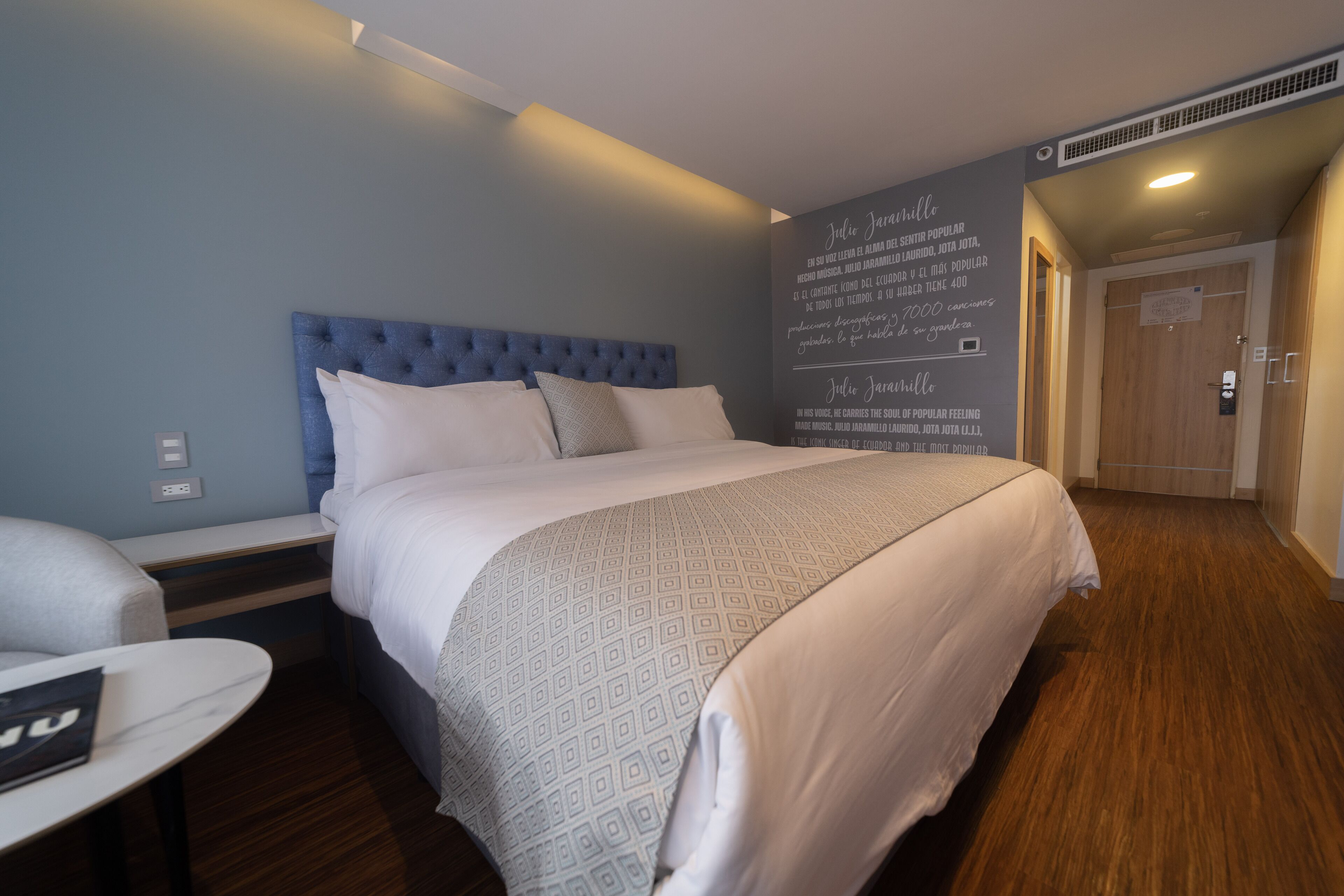 A hotel room with a large bed, a bedside table, a chair, and a wall with a text message.