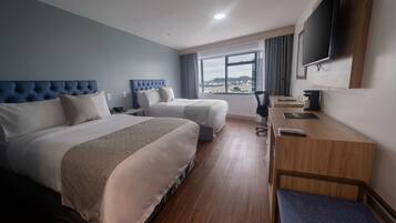 Habitación de hotel con dos camas, un escritorio, un televisor y un amplio ventanal con vistas a la ciudad.