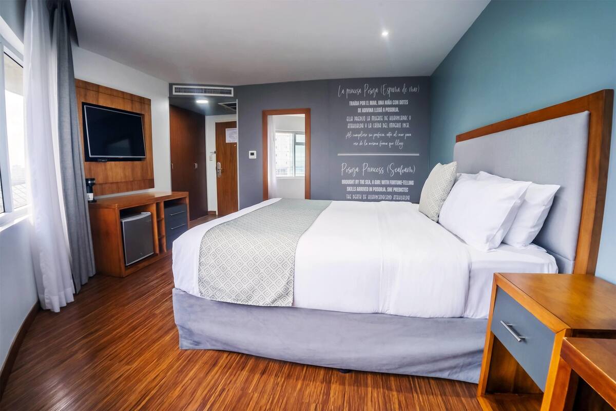 Ein modernes Hotelzimmer mit einem großen Bett, einem an der Wand montierten Fernseher, einem Holztisch und einem kleinen Schrank.