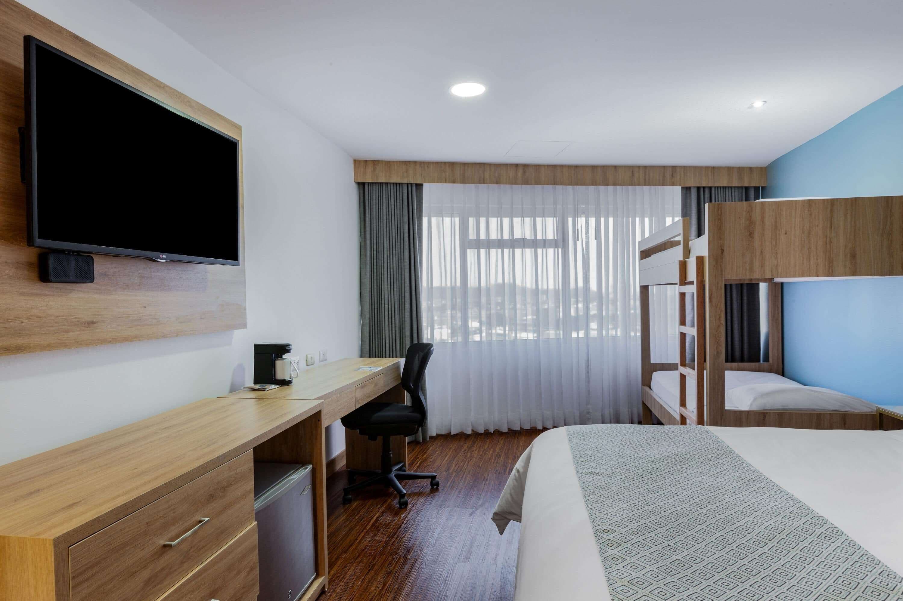 Une chambre d’hôtel avec un bureau en bois, un téléviseur fixé au mur, un lit superposé et une grande fenêtre ornée de rideaux légers.
