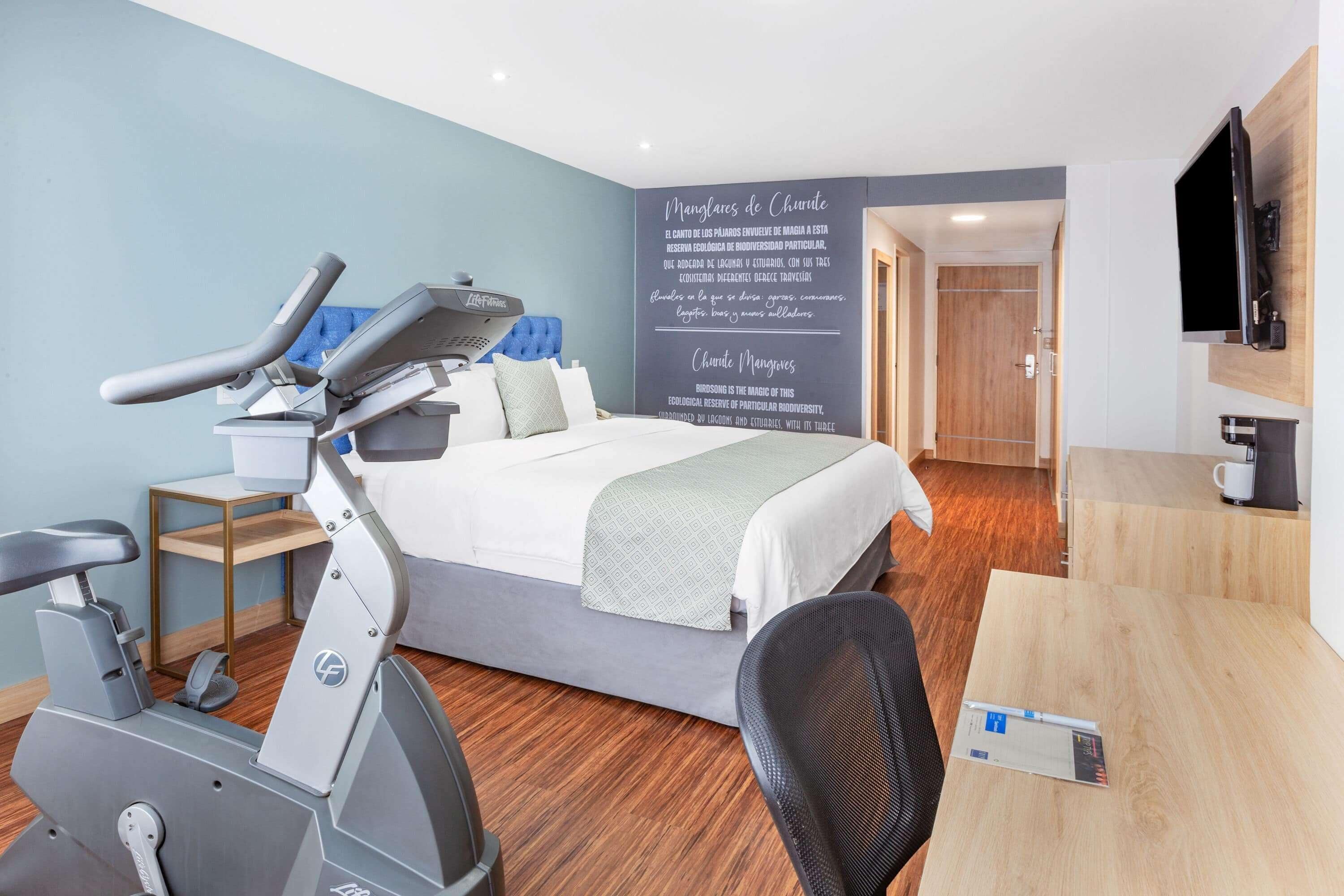 A hotel room with a bed, exercise bike, desk, and a wall with text.