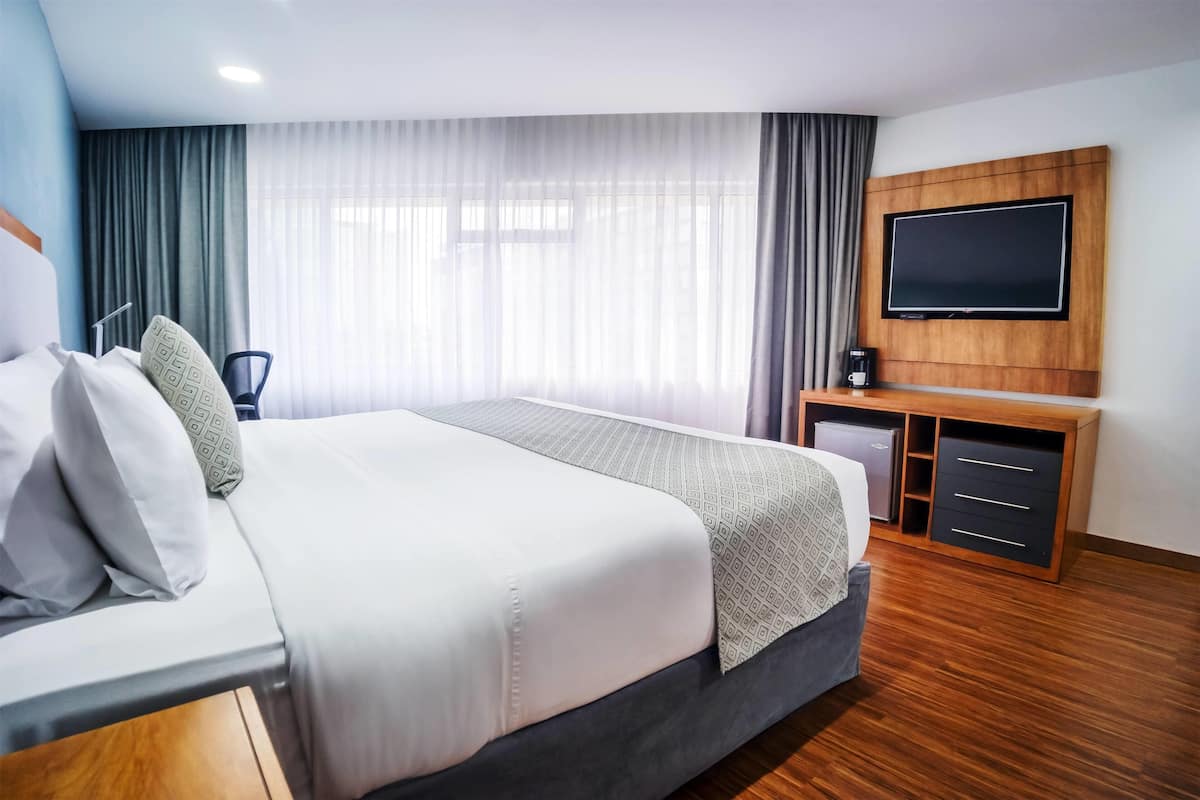Ein modernes Hotelzimmer mit einem großen Bett, einem flachbildfernseher an der Wand, einem Schreibtisch mit Stuhl und einem kleinen Schrank.