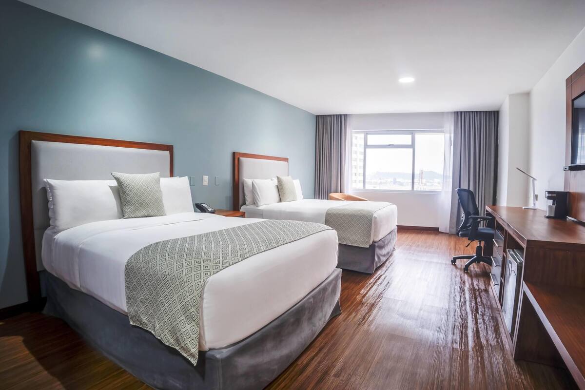 Ein Hotelzimmer mit zwei Betten, einem Schreibtisch, einem Stuhl und einem großen Fenster.