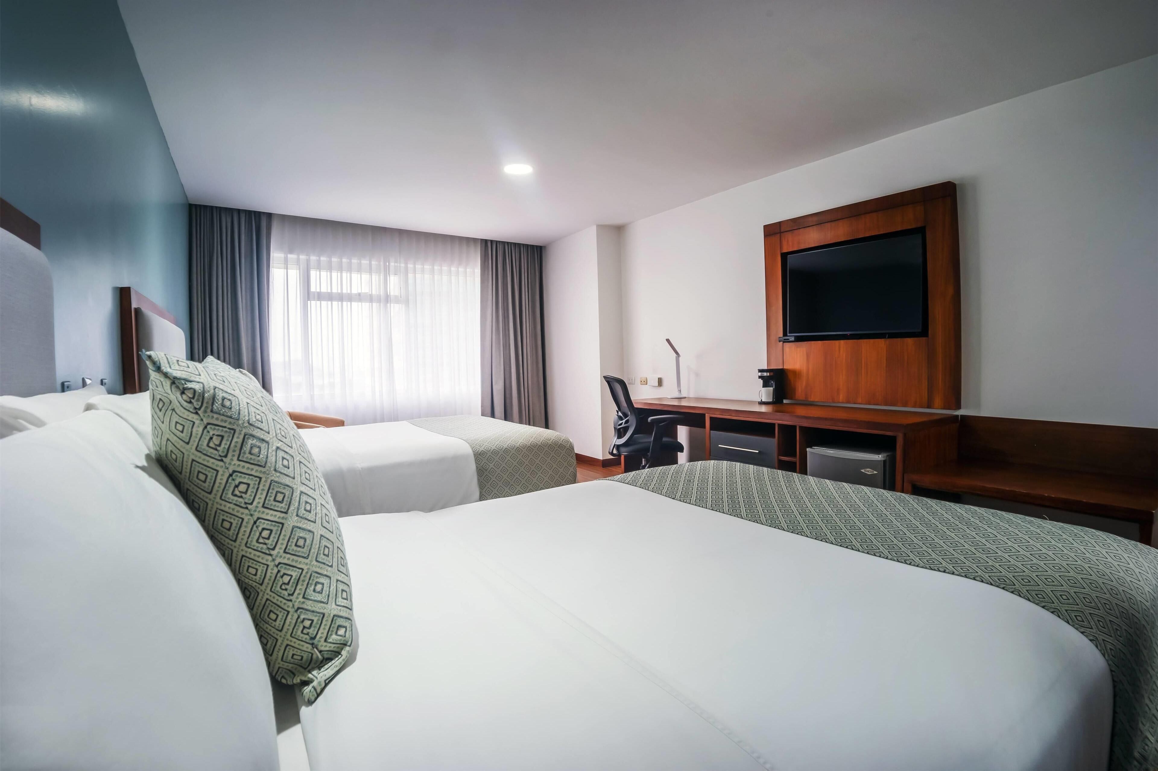 A hotel room with two beds, a desk, a chair, a flat-screen TV, and a window with curtains.