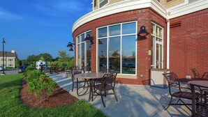 An outdoor patio area with tables and chairs, a brick building with large windows, and a clear blue sky.
