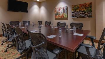 A conference room with a long table, chairs, water bottles, a flat-screen TV, and two paintings on the walls.