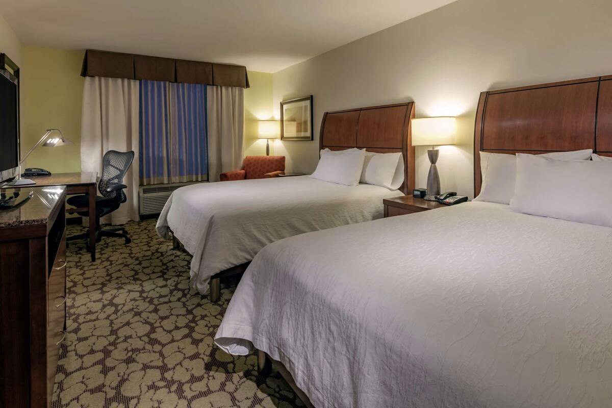 A hotel room with two beds, a desk, a chair, a lamp, and a window with curtains.