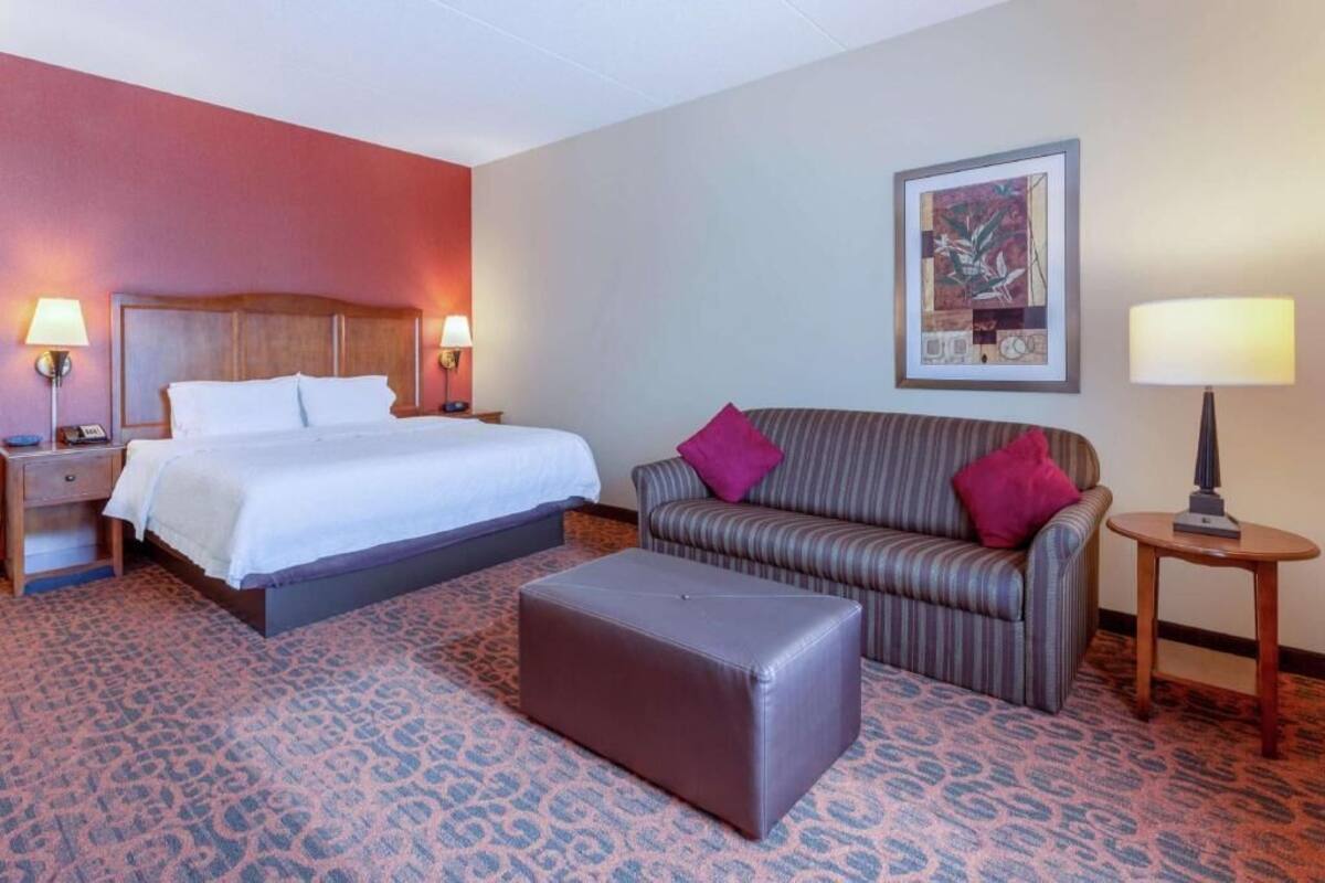 A hotel room with a bed, a sofa, an ottoman, a nightstand, and a lamp.