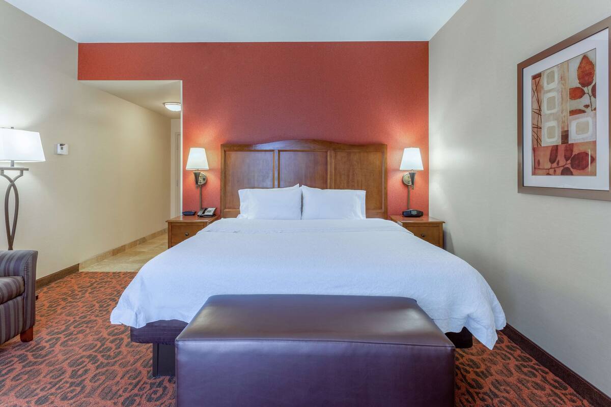 A hotel room with a large bed, a bench, a nightstand with a lamp, and a framed picture on the wall.