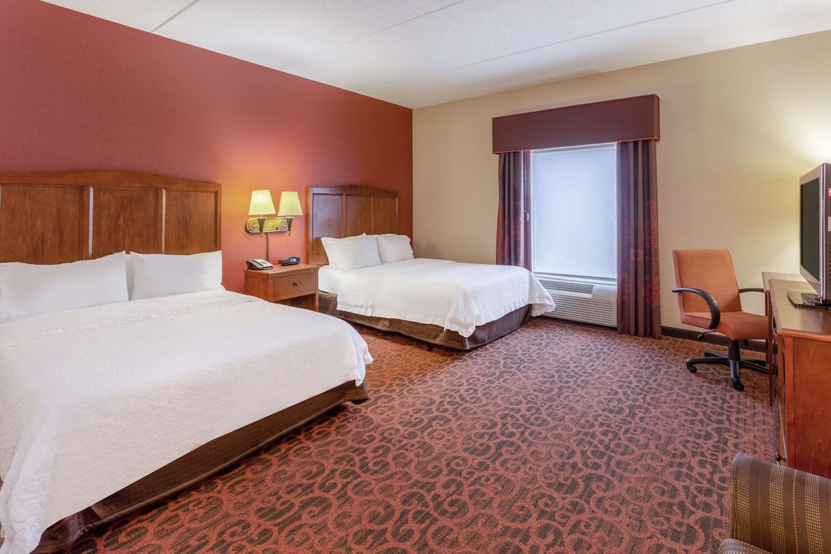 A hotel room with two beds, a desk, a chair, a TV, and a window with curtains.