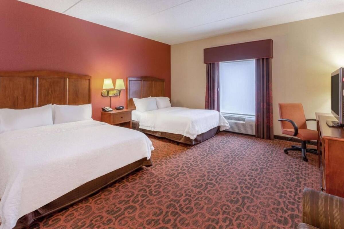 A hotel room with two beds, a desk, a chair, a TV, and a window with curtains.