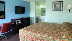 Una habitación de hotel con una cama, un escritorio con computadora y televisor, una silla de oficina roja, un microondas, un baño con espejo y toallero.