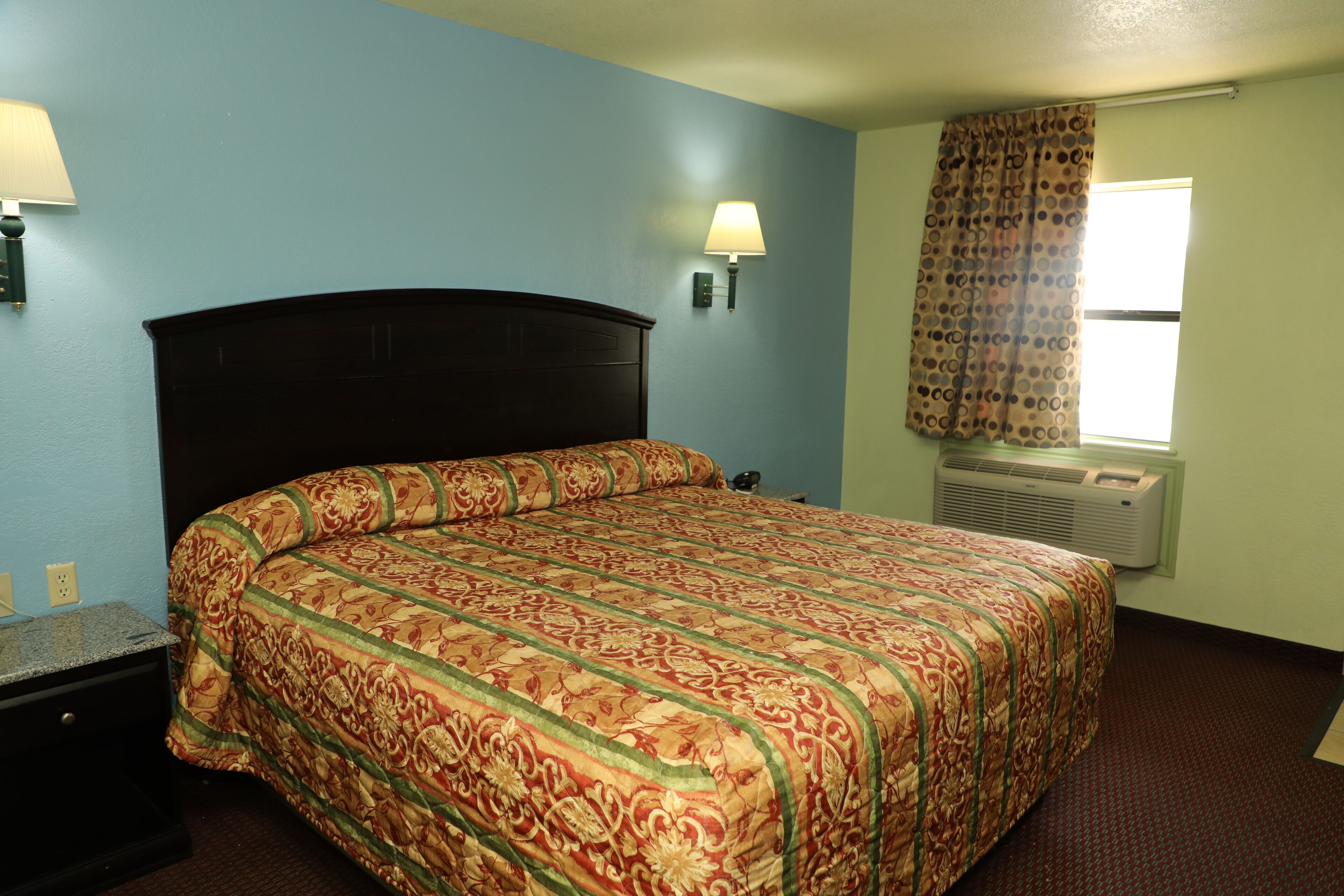 A hotel room with a large bed, two wall-mounted lamps, a window with a curtain, and an air conditioning unit.