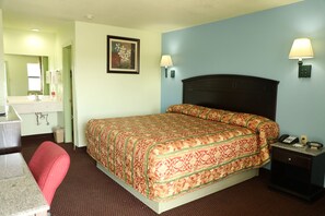 A hotel room with a bed, a desk, a chair, a nightstand, a lamp, and a painting on the wall. - Budget Inn (Stephenville)