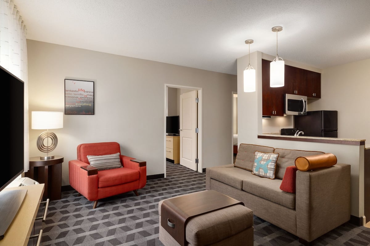 A hotel room with a red armchair, a beige sofa, a small wooden table, a TV, a lamp, a kitchen area with a microwave and refrigerator, and a patterned carpet.