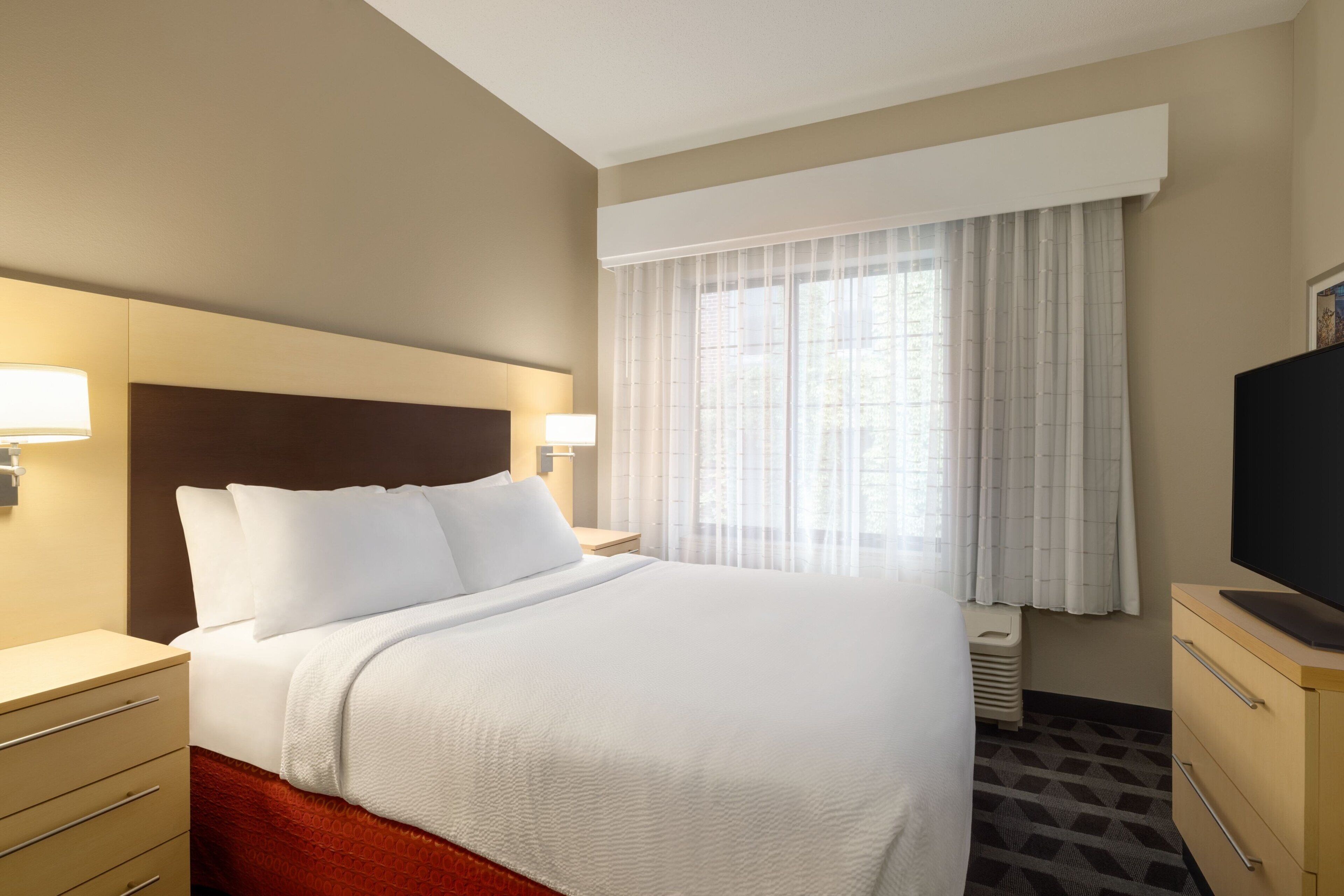 A hotel room with a large bed, bedside lamps, a television, and a window with curtains.