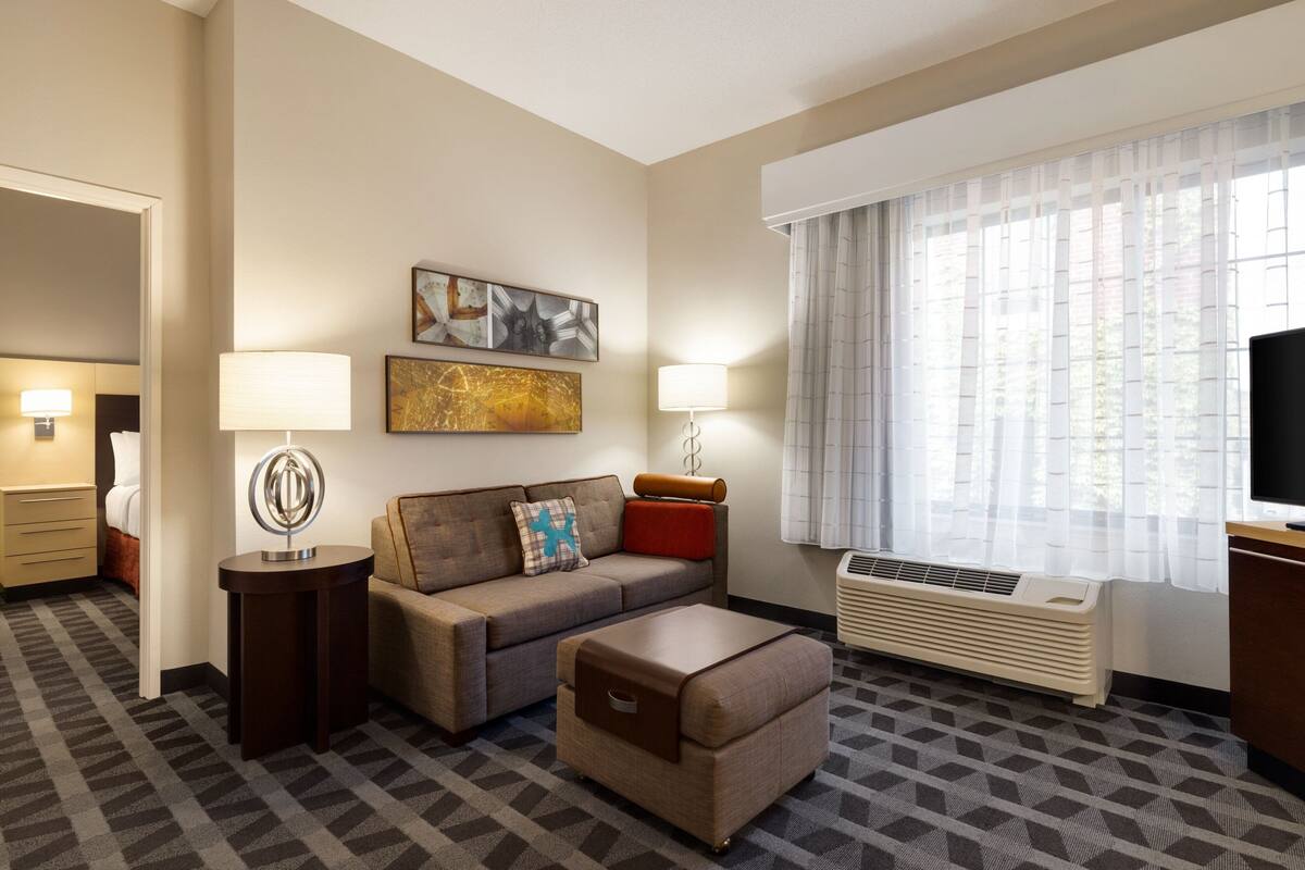 A hotel room with a sofa, ottoman, two bedside tables with lamps, a flat-screen TV, a window with sheer curtains, and a radiator.