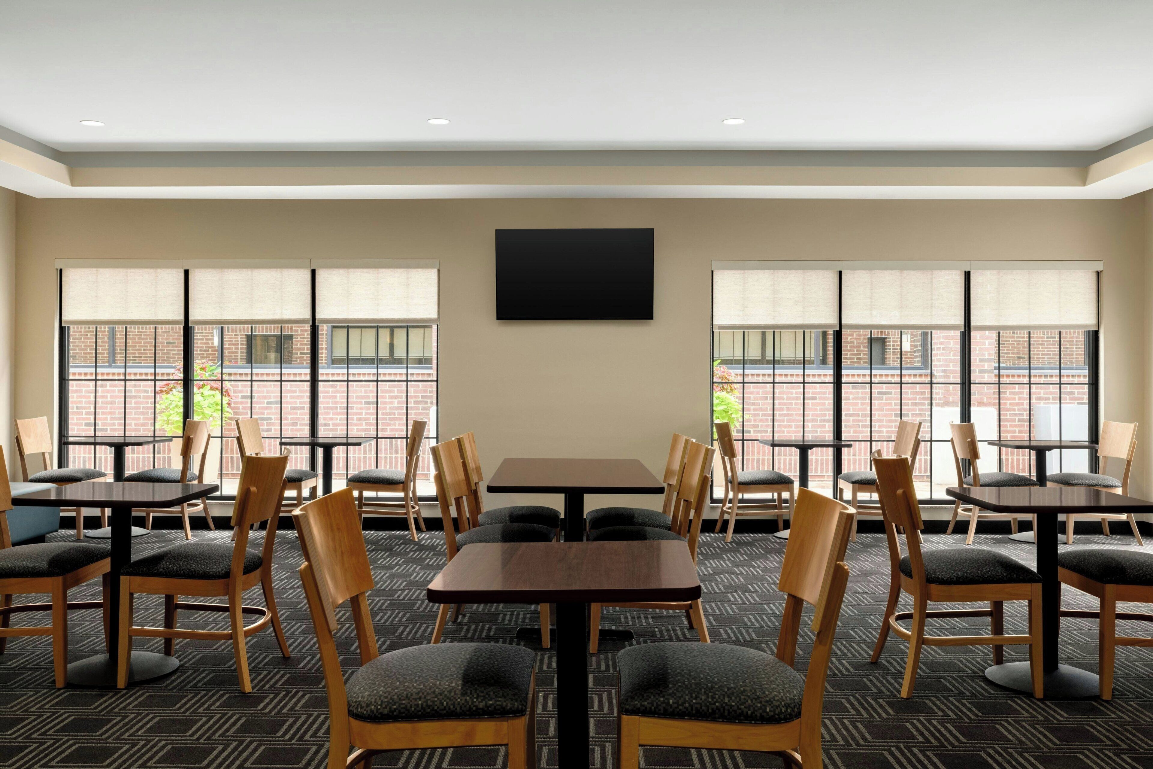 Een vergaderzaal met meerdere tafels en stoelen, grote ramen met zonwering en een flatscreen televisie aan de wand.
