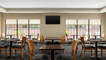 Een vergaderzaal met meerdere tafels en stoelen, grote ramen met zonwering en een flatscreen televisie aan de wand.