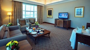 A hotel room with a sofa, armchair, dining table set with plates and glasses, a television on a cabinet, and framed artwork on the walls. - Ayla Hotel  (Al Ain)