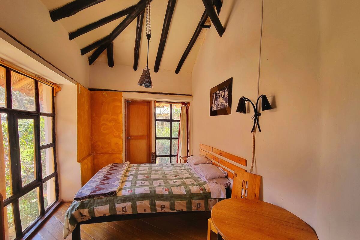 A bedroom with a wooden bed, a small wooden table, and a window with a view of greenery.
