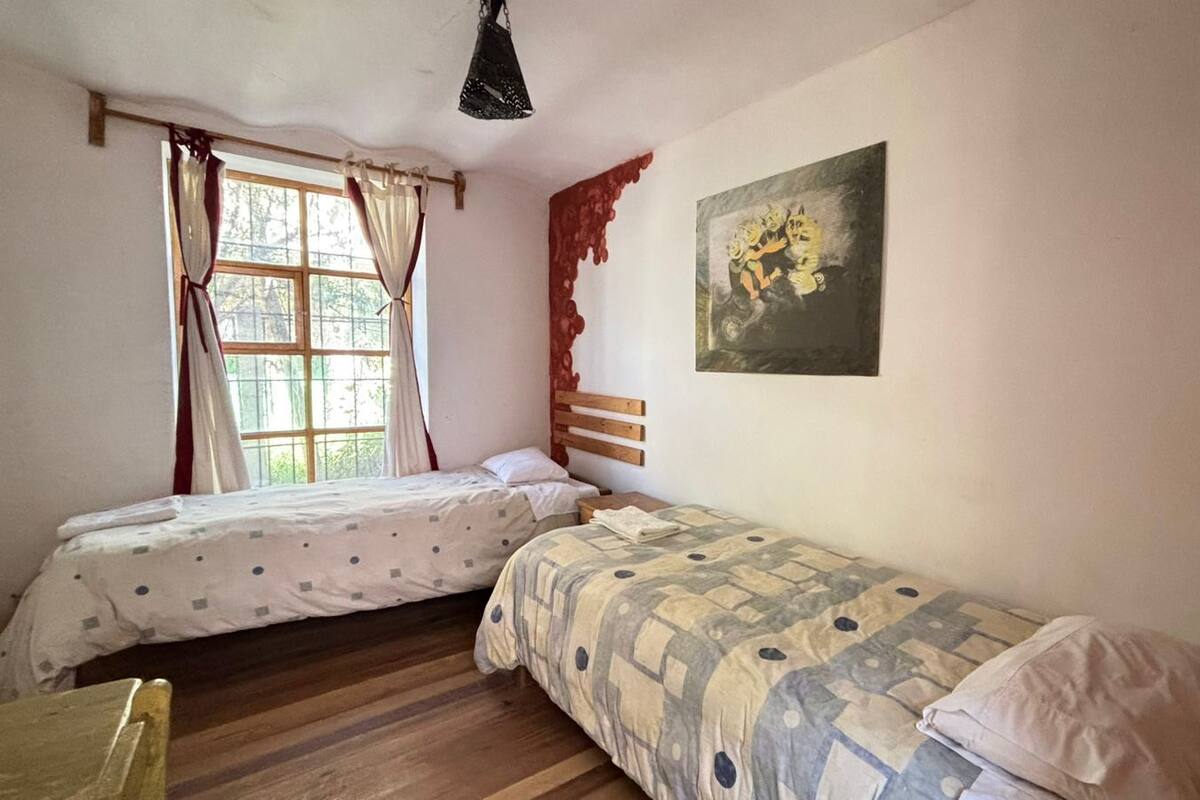 A room with two single beds, a window with curtains, a painting on the wall, and a wooden floor.