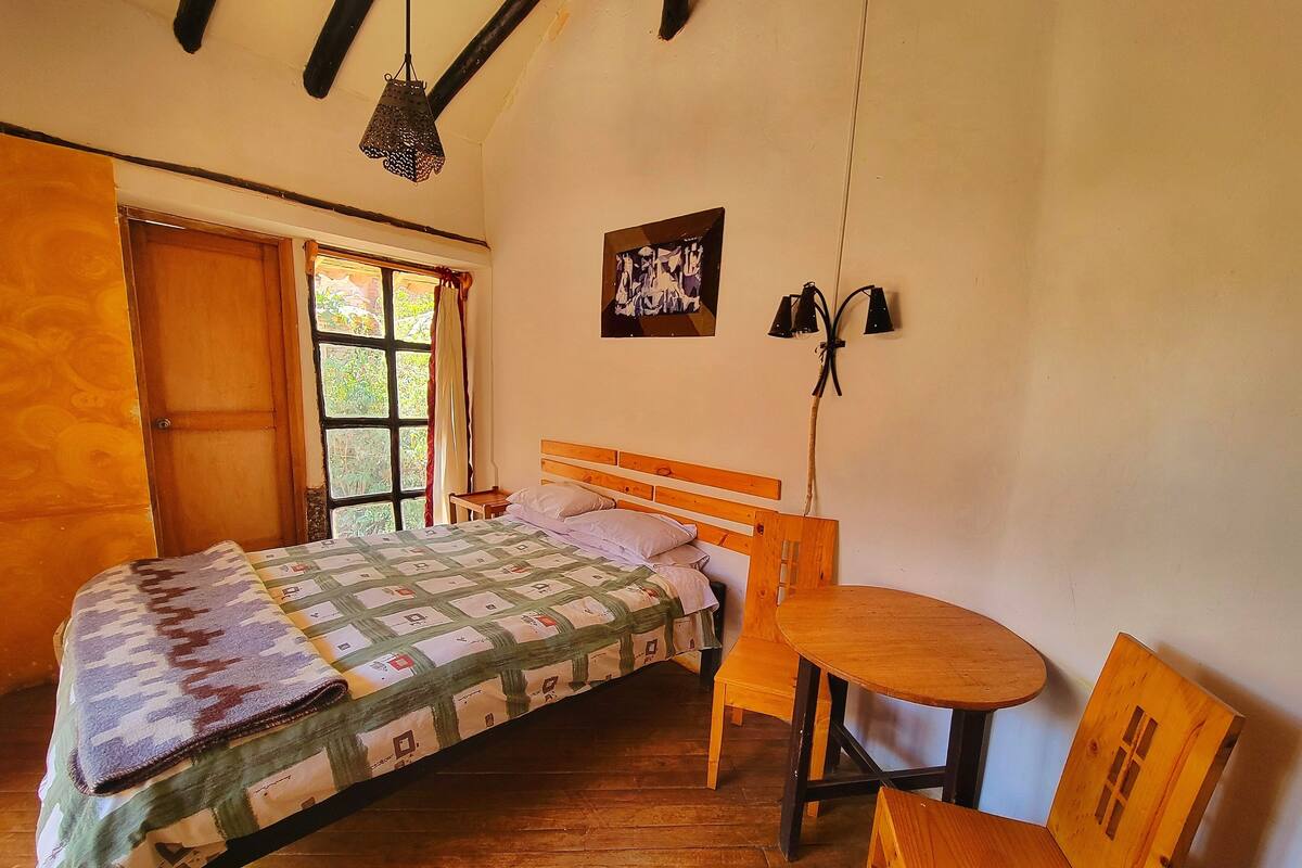 A bedroom with a wooden bed, a small round table, and a chair. There is a window with curtains, a wall-mounted lamp, and a framed picture.