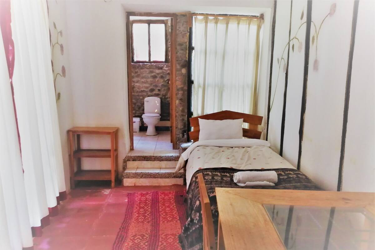 A small, narrow room with a bed, a wooden table, a small stool, a patterned rug, and a view of a bathroom through a doorway.