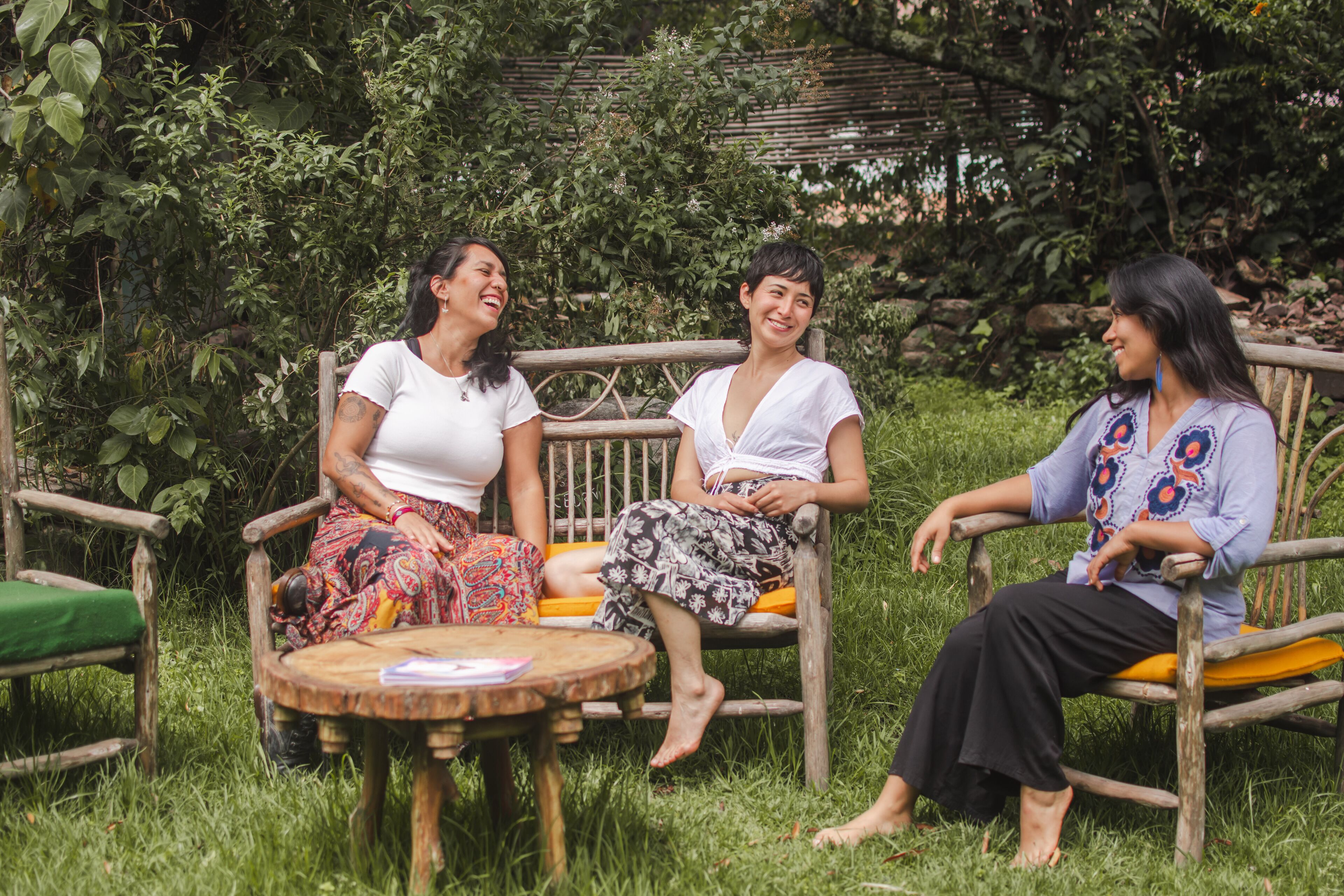Tres personas sentadas en sillas de madera en un jardín, con una mesa redonda de madera al frente.