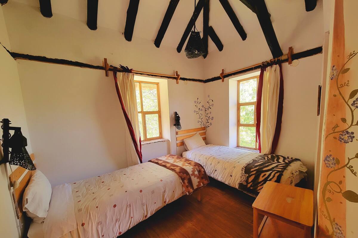 A cozy bedroom with two single beds, wooden flooring, a sloped ceiling with exposed beams, and a window with curtains.