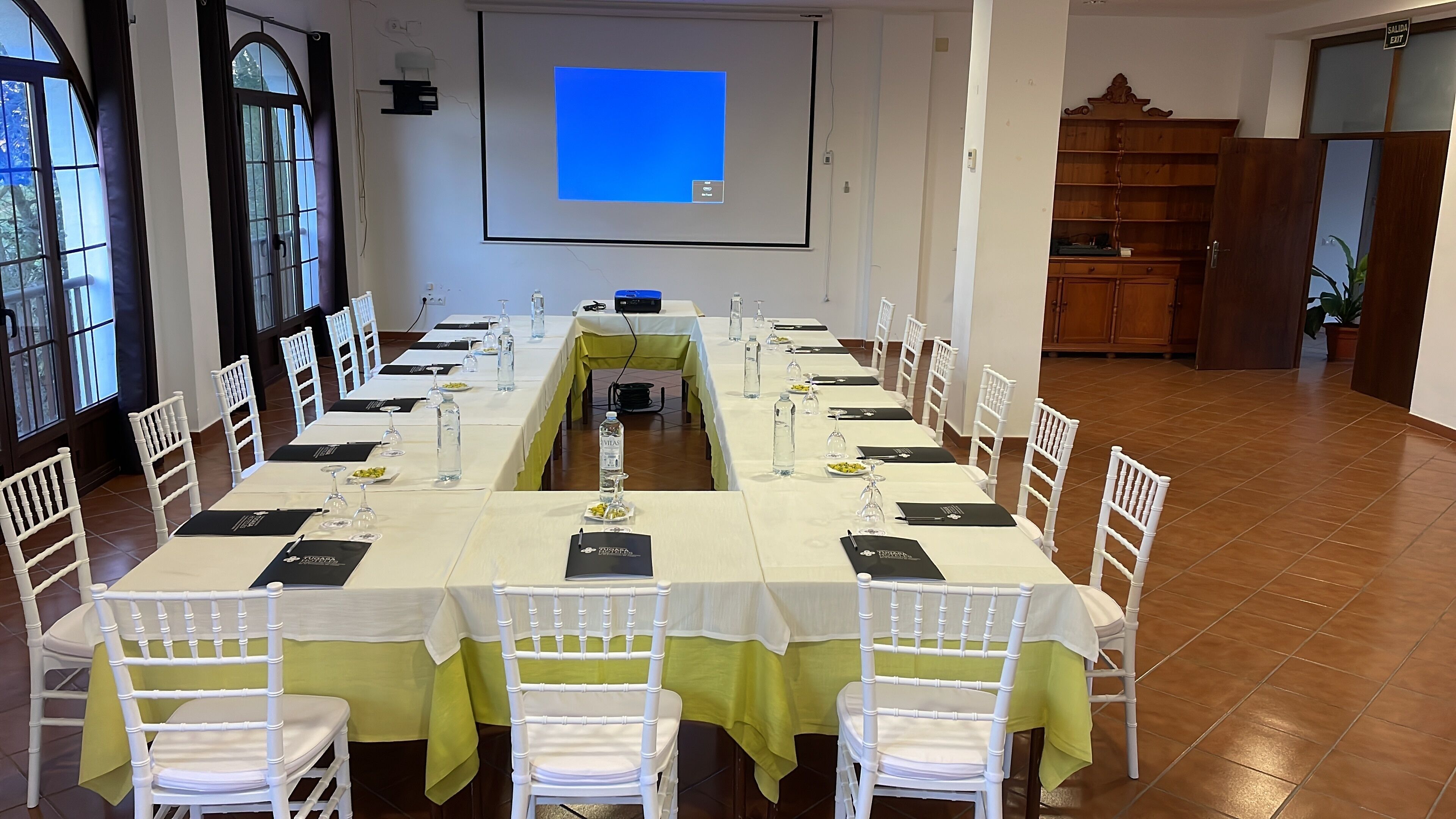 Ein Konferenzraum mit langem Tisch für eine Besprechung, weißen Stühlen, einer Projektionsleinwand und einem Holzschrank im Hintergrund.