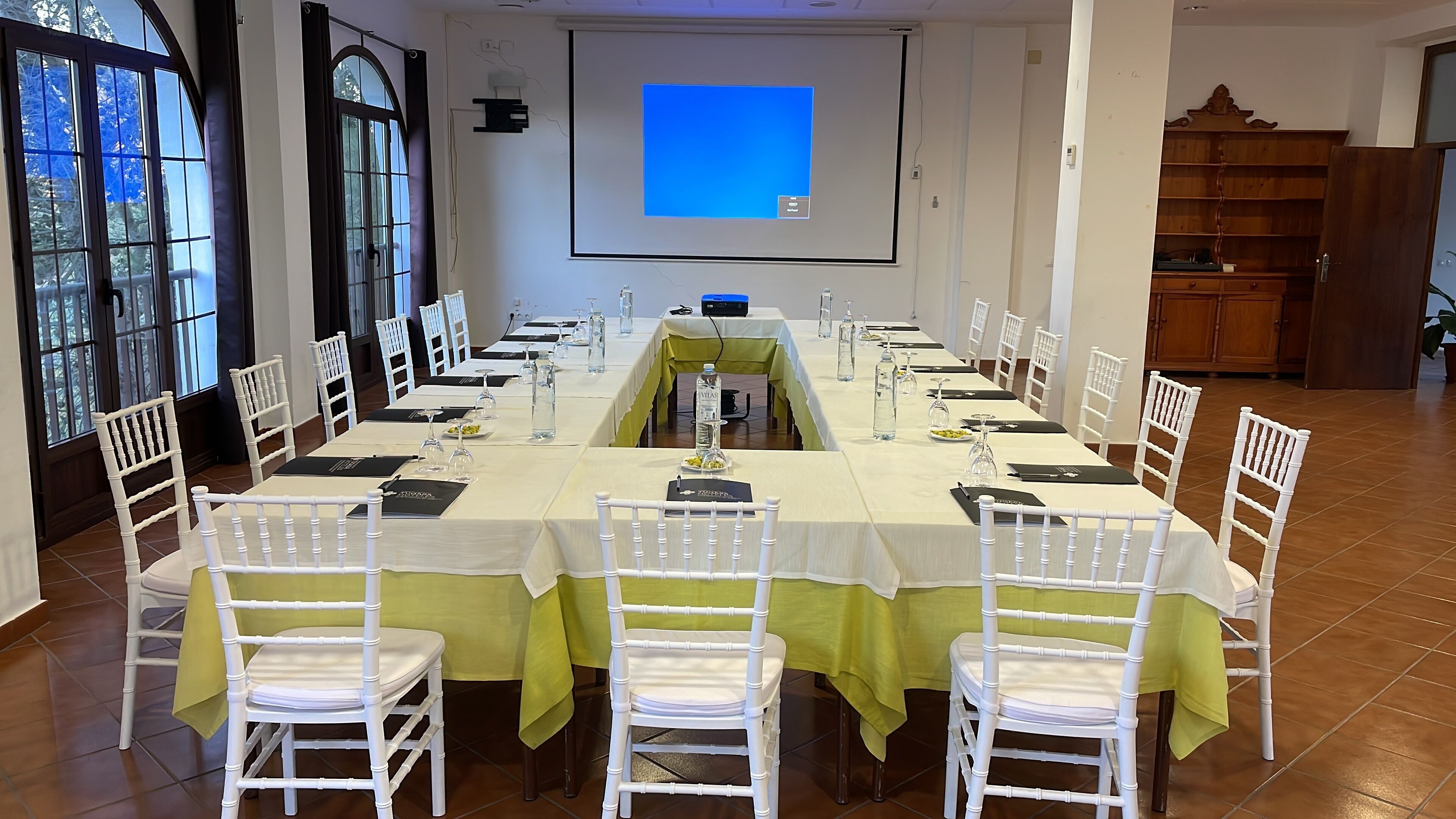 Un salón de conferencias con una larga mesa, sillas blancas y una pantalla para proyector.
