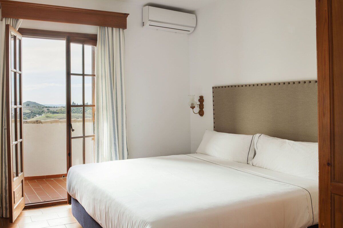 A hotel room with a large bed, a view through a sliding door, and an air conditioning unit on the wall.