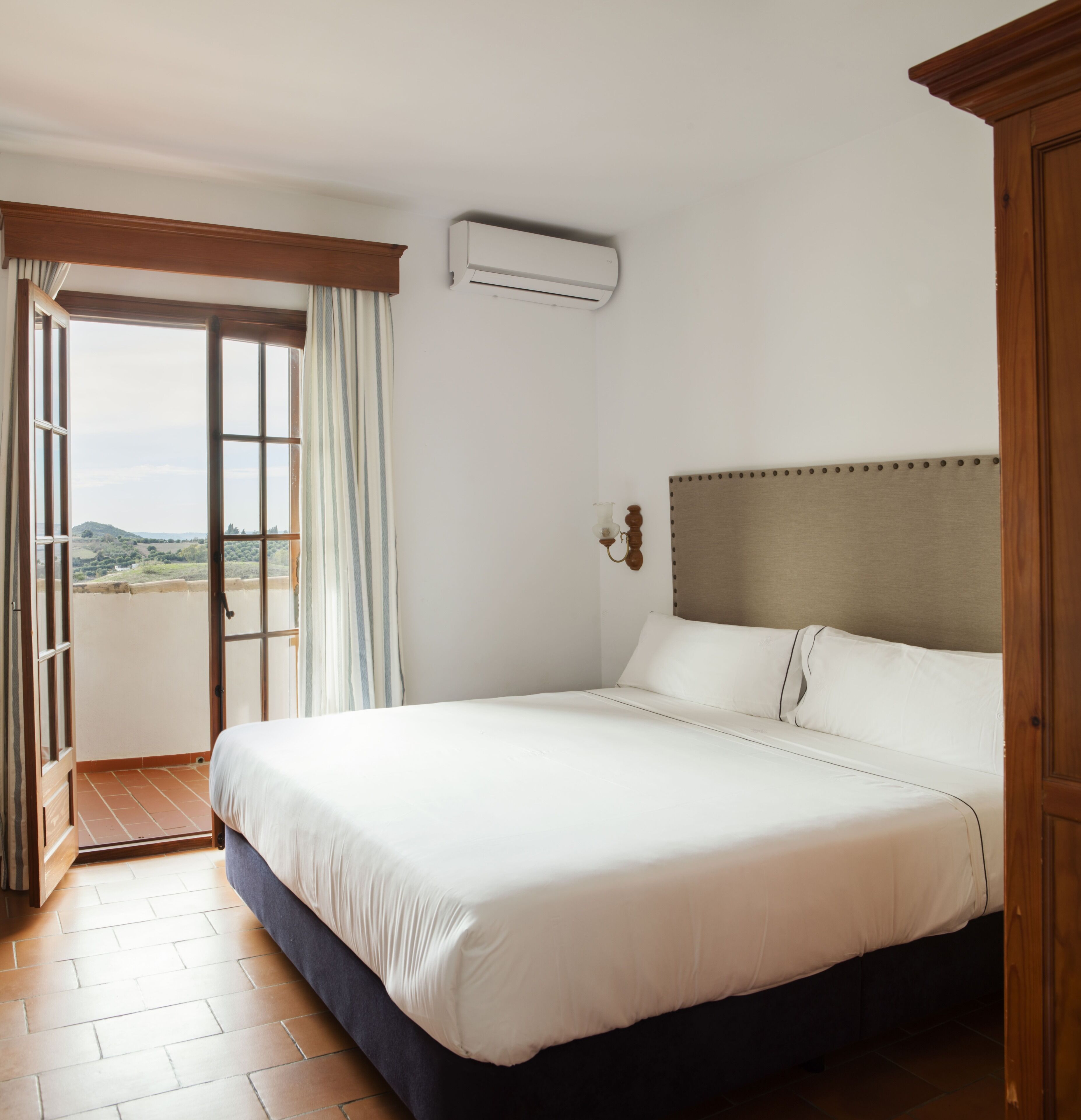 A hotel room with a large bed, a view through an open door, and an air conditioning unit on the wall.