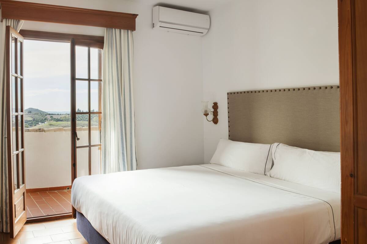 A hotel room with a large bed, a view through an open door, and an air conditioning unit on the wall.