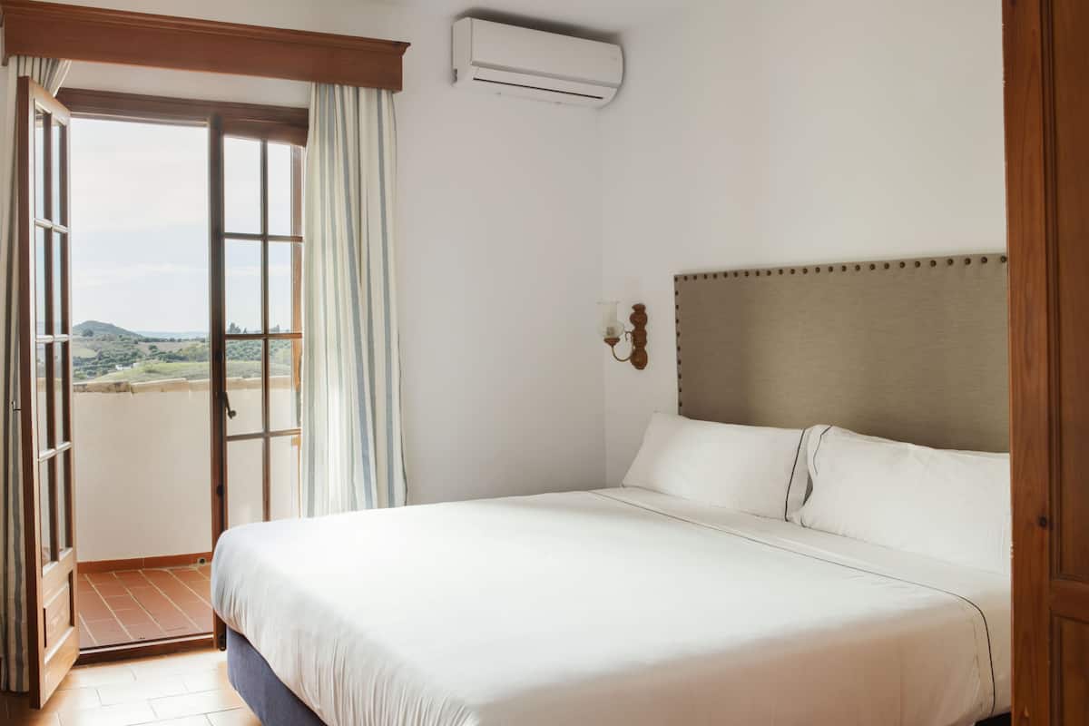 A hotel room with a large bed, a view through an open door, and an air conditioning unit on the wall.