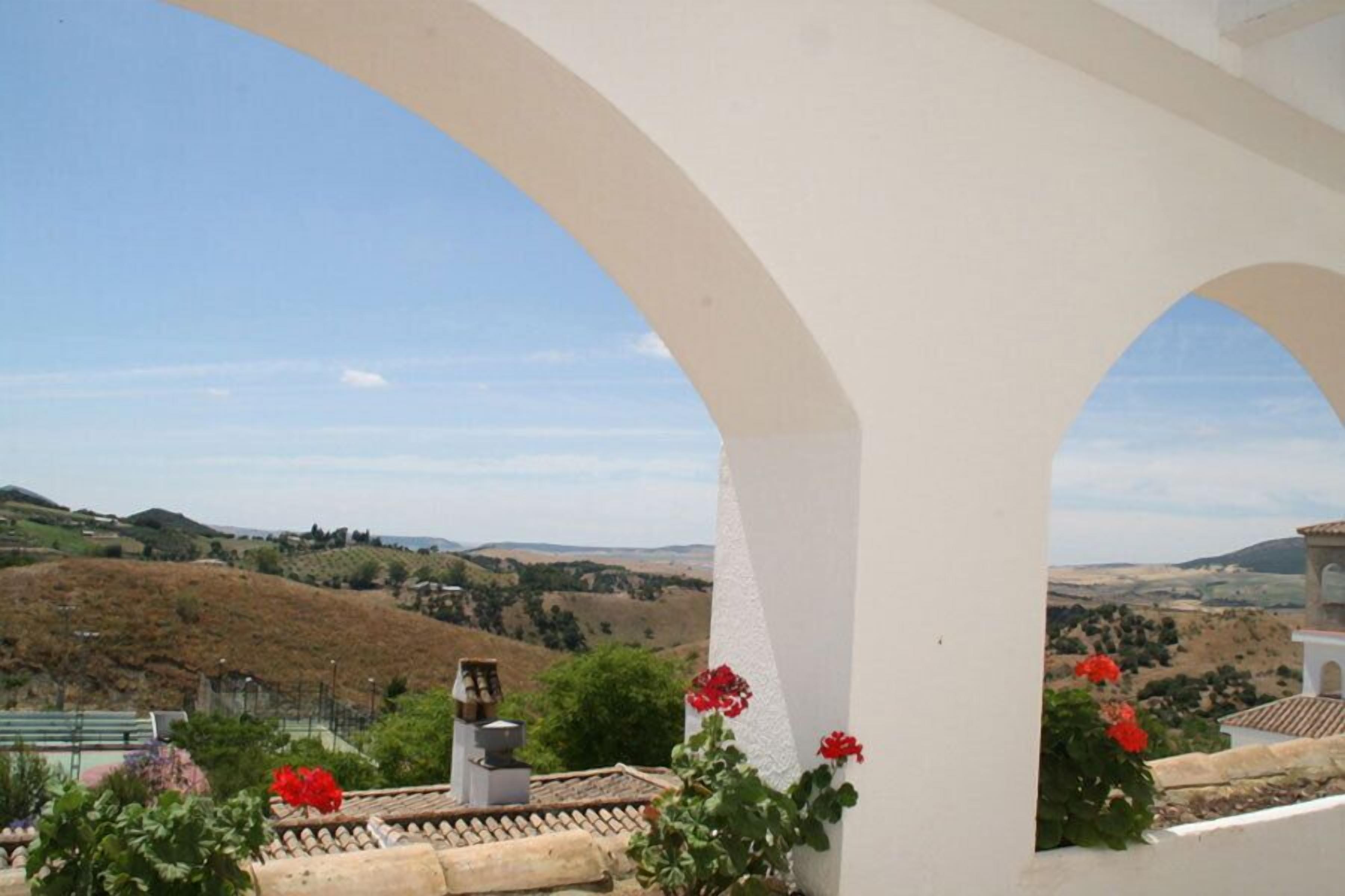 A view from a white-walled balcony with red flowers, overlooking a hilly landscape.