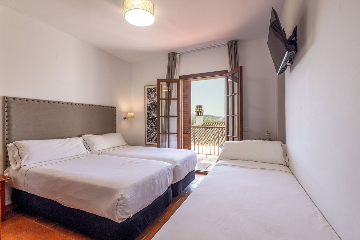 Une chambre d’hôtel avec deux lits, un téléviseur à écran plat fixé au mur et un balcon offrant une vue sur les collines.