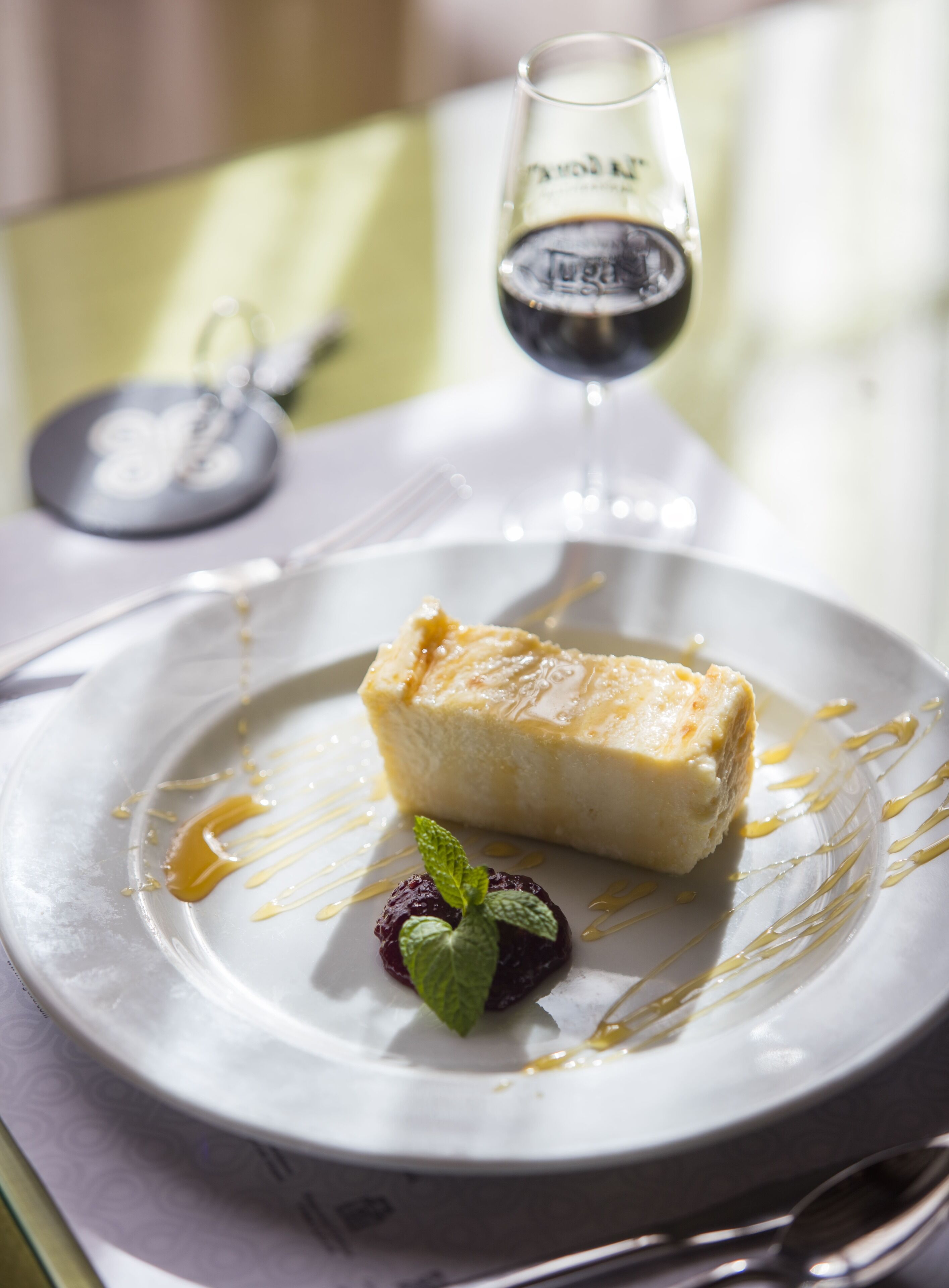 Een dessertborden met een stuk cheesecake, een bessenfruitje en een muntblaadje, vergezeld van een glas rode wijn.