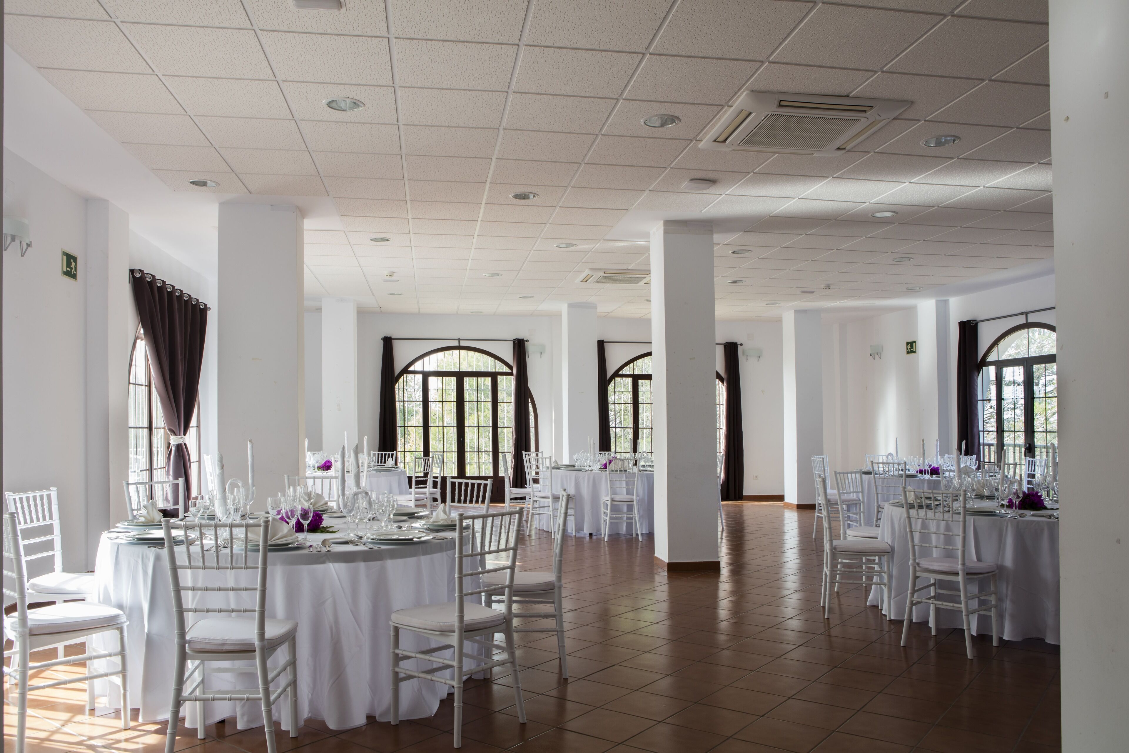 A spacious banquet hall with multiple round tables set for dining, white chairs, and large windows allowing natural light.