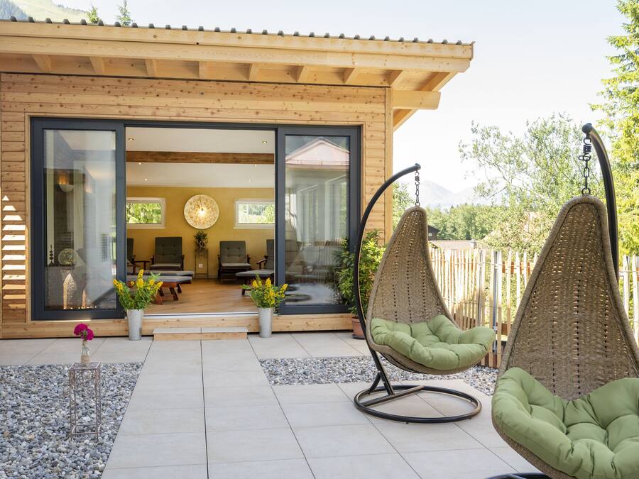 A modern house with a wooden exterior, large glass doors leading to a living area with a sofa and a hanging chair.